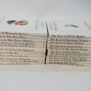 May include: A collection of children's books, including "The Tale of Peter Rabbit" and "The Tale of Benjamin Bunny." The books have white covers with illustrations and text. The books are stacked in two separate piles.