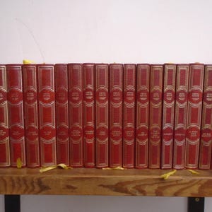 May include: A collection of seventeen red hardcover books arranged in a row. Each book spine features gold-colored lettering and decorative gold-colored lines. The books are positioned upright, displaying their spines with uniform height and design.