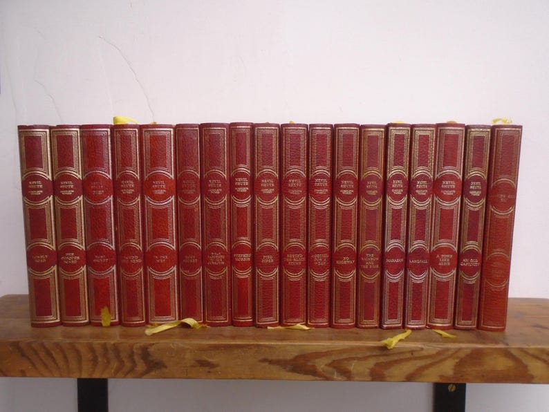 May include: A collection of seventeen red hardcover books arranged in a row. Each book spine features gold-colored lettering and decorative gold-colored lines. The books are positioned upright, displaying their spines with uniform height and design.