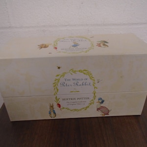 Puede incluir: Una caja blanca con una tapa de color crema. La caja tiene una imagen de Peter Rabbit en la parte delantera y el texto "The World of Peter Rabbit" y "Beatrix Potter - The Complete Collection of Original Tales 1-23".