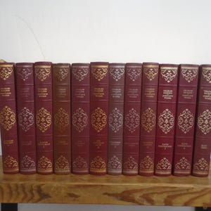 Op de afbeelding: Een collectie van twaalf boeken met harde kaft van Charles Dickens, voorzien van dieprode ruggen met gedetailleerde gouden filigraanpatronen en belettering. Elke rug toont de naam van de auteur en de titel in een uniforme, decoratieve stijl.