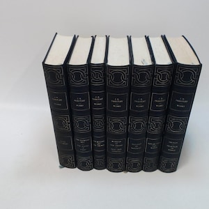 May include: A collection of seven hardcover books with dark blue covers and gold lettering. The books are arranged upright, with their spines facing the viewer. The titles and author's name are visible on the spines. The pages are white.