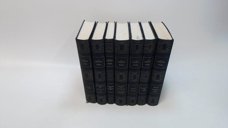 May include: A collection of seven hardcover books with dark blue covers and gold lettering. The books are arranged upright, with their spines facing the viewer. The titles and author's name are visible on the spines. The pages are white.