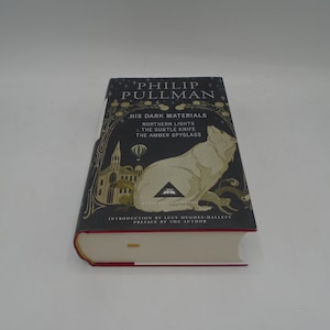 Peut inclure: Un livre de Philip Pullman intitulé "His Dark Materials". La couverture sombre présente une illustration blanche d'une créature ressemblant à un loup. La tranche du livre est rouge, et les pages sont blanches.