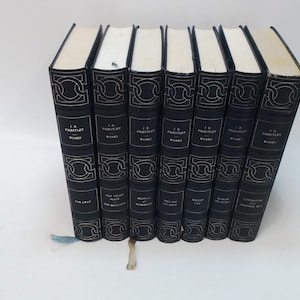May include: A collection of seven hardcover books with black covers and gold decorative accents. The spines feature the author's name, title, and series information. The pages are cream-colored, and a blue ribbon bookmark is visible.