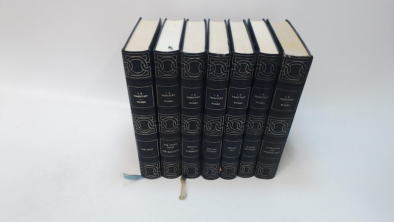 May include: A collection of seven hardcover books with black covers and gold decorative accents. The spines feature the author's name, title, and series information. The pages are cream-colored, and a blue ribbon bookmark is visible.