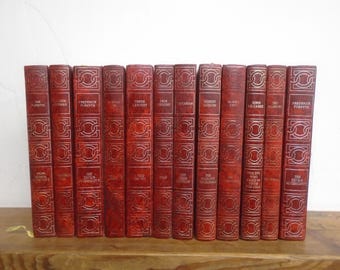 Puede incluir: Una colección de doce libros encuadernados en cuero rojo dispuestos verticalmente en fila. Cada lomo presenta patrones geométricos en relieve de color dorado y letras que indican el autor y el título de la obra, creando un aspecto uniforme.