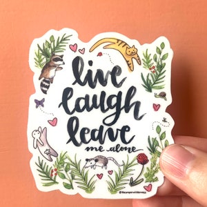 May include: A white sticker with the words "live laugh leave me alone" in black script. The sticker features illustrations of a raccoon, cat, opossum, and other small creatures, surrounded by greenery and hearts.