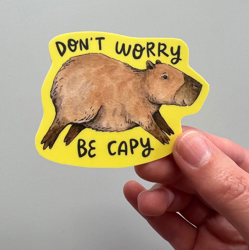 May include: A yellow sticker with the text "DON'T WORRY BE CAPY" and a watercolor illustration of a capybara. The capybara is brown with a dark brown nose and feet. The sticker is held by a hand.