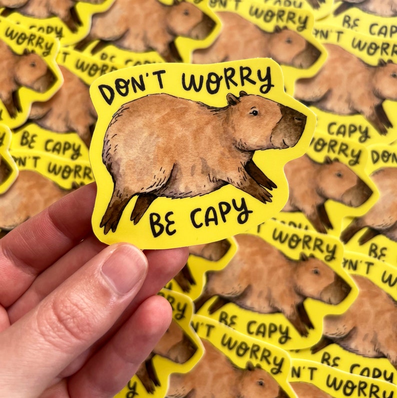 May include: A yellow sticker with a watercolor illustration of a capybara. The sticker reads "DON'T WORRY BE CAPY" in black text. The capybara is brown with a dark brown face and legs. The sticker is held by a hand.