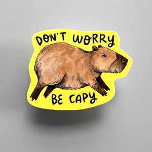 May include: A yellow sticker featuring a watercolor illustration of a capybara. The capybara is brown with a dark brown face and legs. The text "DON'T WORRY BE CAPY" is written in black above and below the capybara.