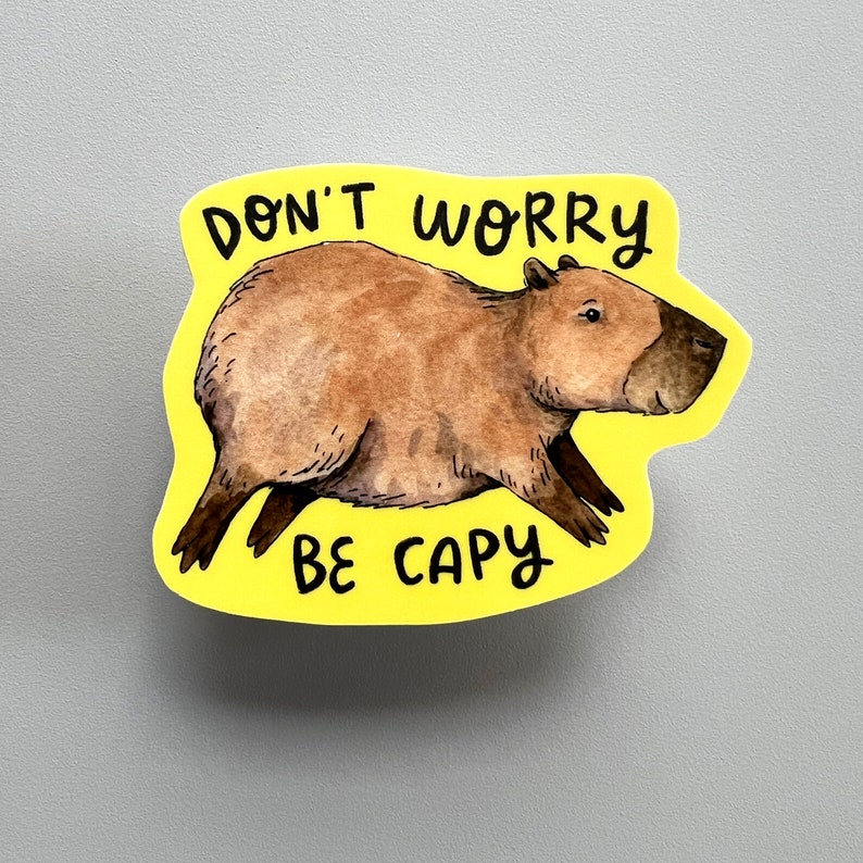 May include: A yellow sticker featuring a watercolor illustration of a capybara. The capybara is brown with a dark brown face and legs. The text "DON'T WORRY BE CAPY" is written in black above and below the capybara.