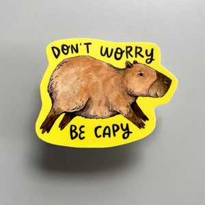 May include: A yellow sticker with a watercolor illustration of a capybara. The capybara is brown with a dark brown face and legs. The sticker has the text "DON'T WORRY BE CAPY" in black.