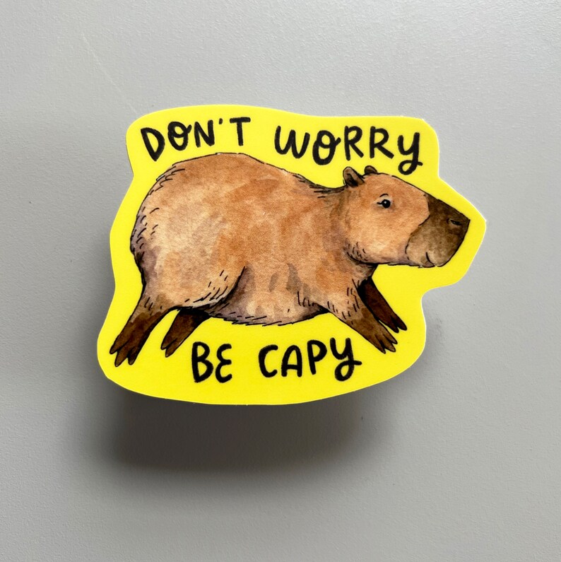 May include: A yellow sticker with a watercolor illustration of a capybara. The capybara is brown with a dark brown face and legs. The sticker has the text "DON'T WORRY BE CAPY" in black.
