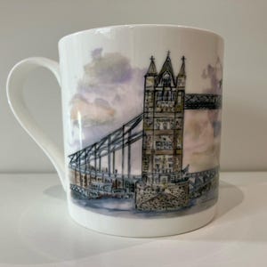 Pode incluir: Caneca de cerâmica branca com uma ilustração em aquarela da Tower Bridge em Londres. O design inclui as torres da ponte, as passarelas e o rio, contra um céu nublado. A caneca tem uma alça curva.
