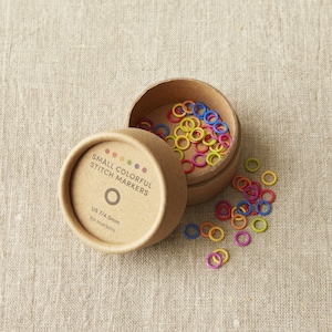 May include: A round cardboard box with a lid containing colourful stitch markers. The box is labelled "Small Colorful Stitch Markers" and "US 7/4.5mm 60 markers".