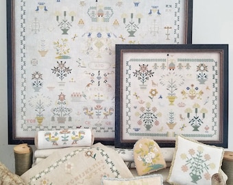 May include: A collection of cross-stitch embroidery projects featuring floral and bird motifs. The projects are framed and displayed on a table with a variety of colors, including pink, blue, green, and yellow. The projects include a sampler with the alphabet, a heart-shaped pillow, and a square pillow.