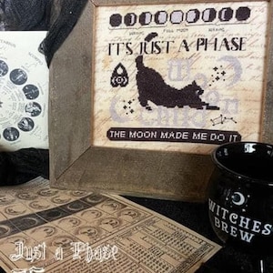 May include: A cross-stitch pattern featuring a black cat and the text "It's Just a Phase" and "The Moon Made Me Do It". The pattern is framed in a rustic wooden frame.