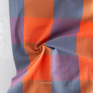 May include: A close-up of a fabric with a large plaid pattern in shades of orange, blue, and purple. The fabric is folded, showing texture and depth. The word "FABLEISM" is printed on the fabric.