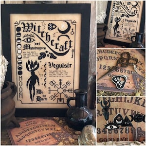 May include: A framed cross-stitch pattern featuring the words "Witchcraft" and "Veguisir" in black lettering on a beige background. The design includes a moon, stars, and a stylised plant with the words "Mandragola" and "Do No Harm".