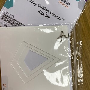 Könnte beinhalten: Ein weißer, plastik-geformter Drachen-Betrachter mit einer Kette. Der Betrachter ist in einer durchsichtigen Plastiktüte verpackt, mit einer weißen Karte, auf der "Fussy Cutting Viewers™ Kite Set" und "Imprezzio Notions" steht. Die Karte hat auch einen Barcode und den Text "FCV.KITE" und "www.imprezzio.com".