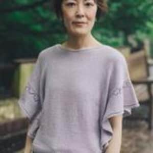 May include: A light purple knit top with short, flared sleeves. The sleeves have a delicate, openwork design near the hem. The top has a round neckline and a relaxed fit, suitable for casual wear. The background is blurred, suggesting an outdoor setting.