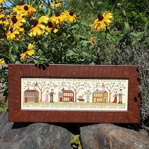 May include: A framed cross-stitch sampler with a brown wooden frame. The sampler depicts a row of houses with trees and birds. The text on the sampler reads "Behold how good & Pleasant it is For Brethren to dwell Together in Unity".