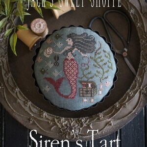 May include: A cross-stitch pattern of a mermaid in a pie crust, with the text "Siren's Tart" and "Original Designs by Paulette Stewart" below. The pattern is in a wooden frame with a green leafy plant in the background.