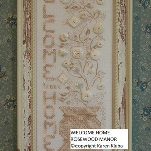 May include: Framed cross-stitch art with the words "WELCOME HOME" and a floral design made of buttons. The artwork is in a distressed cream-coloured frame. The text "WELCOME HOME ROSEWOOD MANOR" is also visible.