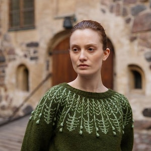 May include: A woman wearing a dark green jumper with a light green embroidered design. The jumper has a crew neck and long sleeves.