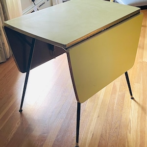 May include: A rectangular drop-leaf table with a light yellow laminate top and matching side leaves. The table features four slender, tapered black metal legs with metallic caps at the base. The structure includes a dark brown support frame.