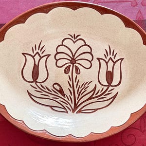 May include: A white ceramic oval serving dish with a brown rim and a floral design. The design features three stylized tulips and a flower with a central stem.