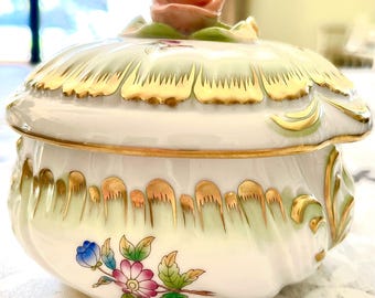 May include: A white porcelain trinket box with a green and gold floral design. The lid features a pink rose with green leaves.