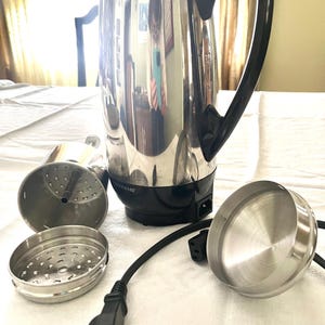 May include: A stainless steel electric coffee percolator with a black handle and base. The disassembled parts, including the filter basket and lid, are visible. The power cord is plugged in.
