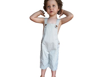 Könnte beinhalten: Ein Kind in einer hellblau-weiß gestreiften Latzhose mit Holzknöpfen. Die Latzhose hat ein klassisches Design mit verstellbaren Trägern und Taschen. Das Kind steht mit erhobenen Armen da.