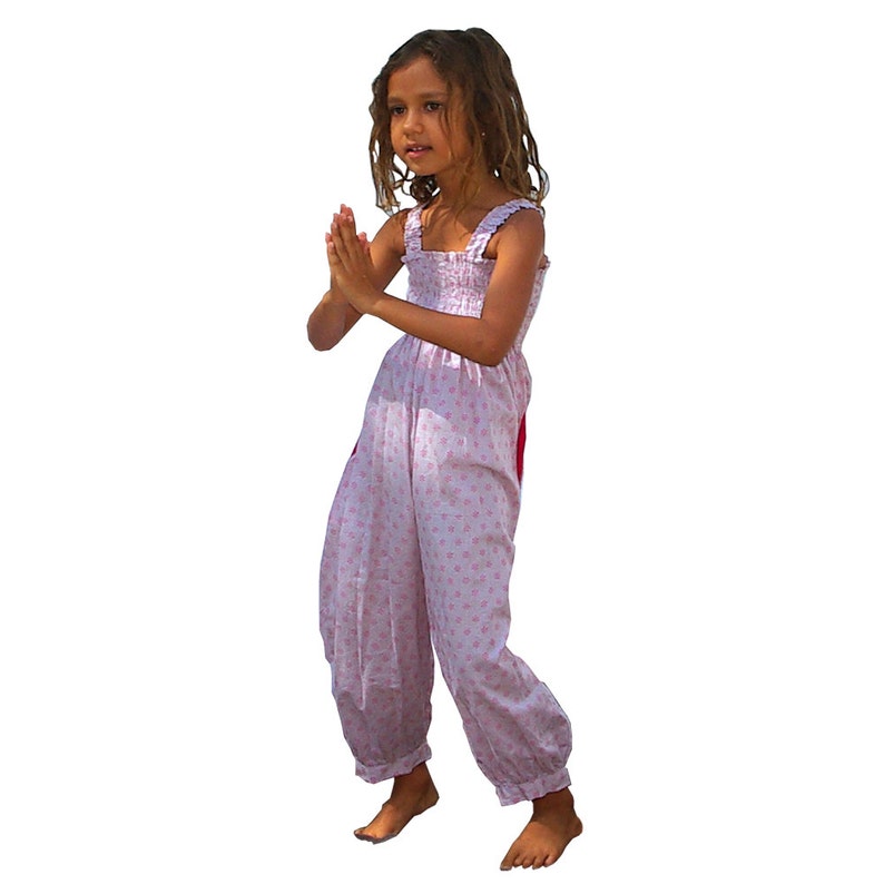 May include: A young girl wearing a pink and white floral print jumpsuit with a red accent. She is standing with her hands clasped in front of her.