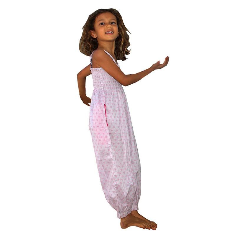 May include: A young girl wearing a white and pink floral print jumpsuit with a pink pocket. She is standing with her right arm raised and her left hand on her hip.