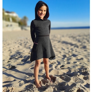 Può includere: Una giovane persona indossa un vestito nero con cappuccio e un motivo geometrico bianco e nero. Il vestito ha una cintura nera e un bordo rosa sul cappuccio. La persona è in piedi su una spiaggia sabbiosa.