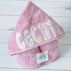 May include: Pink hooded towel with the name "Cecilia" embroidered in colorful floral fabric. The towel is folded and has a small tag with the text "DANDELION CREEK EST 2018".