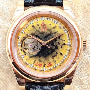 May include: A gold wristwatch with a black leather strap. The watch face has a yellow and black speckled pattern, with a visible mechanical movement and rose gold hands. The face is decorated with small gemstones and the numbers "J.M. 36".