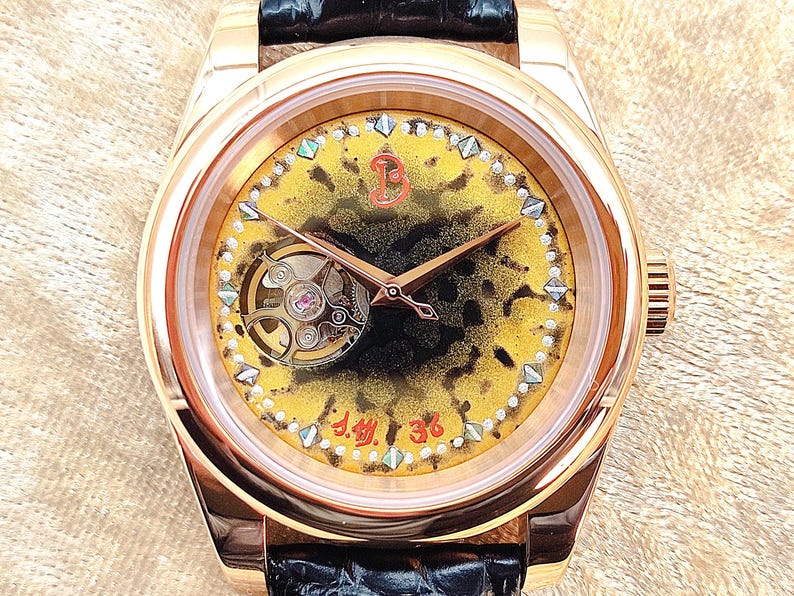 May include: A gold wristwatch with a black leather strap. The watch face has a yellow and black speckled pattern, with a visible mechanical movement and rose gold hands. The face is decorated with small gemstones and the numbers "J.M. 36".