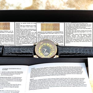 May include: A gold-plated Bottegando watch with a black leather strap. The watch has a distinctive face design and is displayed in a black box with paperwork. The watch has a 40 mm case diameter.