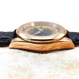 May include: A rose gold wristwatch with a black leather strap. The watch face features a floral design and gold hands. The watch is set against a neutral background, highlighting its elegant design.