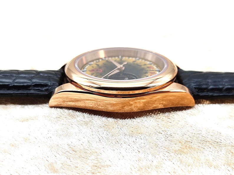 May include: A rose gold wristwatch with a black leather strap. The watch face features a floral design and gold hands. The watch is set against a neutral background, highlighting its elegant design.
