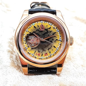 May include: A rose gold wristwatch with a black leather strap. The watch face has a yellow and black patterned design and a visible mechanical movement. The bezel is decorated with small, iridescent accents. The text "J.M. 36" is visible.