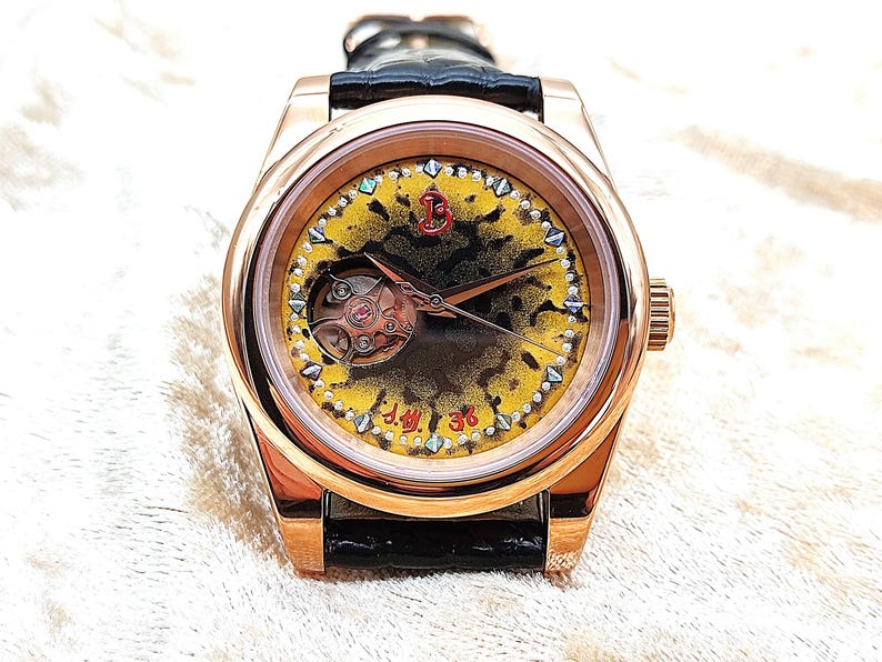 May include: A rose gold wristwatch with a black leather strap. The watch face has a yellow and black patterned design and a visible mechanical movement. The bezel is decorated with small, iridescent accents. The text "J.M. 36" is visible.
