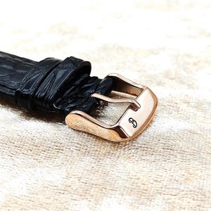 May include: Close-up of a black leather watch strap with a rose gold buckle. The buckle has a small, engraved design. The leather has a textured, crocodile-like pattern.