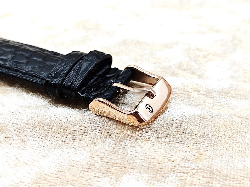 May include: Close-up of a black leather watch strap with a rose gold buckle. The buckle has a small, engraved design. The leather has a textured, crocodile-like pattern.