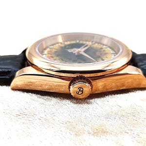 May include: A rose gold wristwatch with a black leather strap. The watch face features a floral design, gold hands, and a gold bezel. The watch crown is engraved with the letter "B".