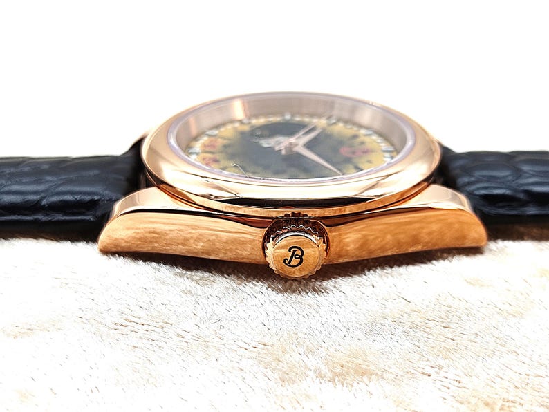 May include: A rose gold wristwatch with a black leather strap. The watch face features a floral design, gold hands, and a gold bezel. The watch crown is engraved with the letter "B".